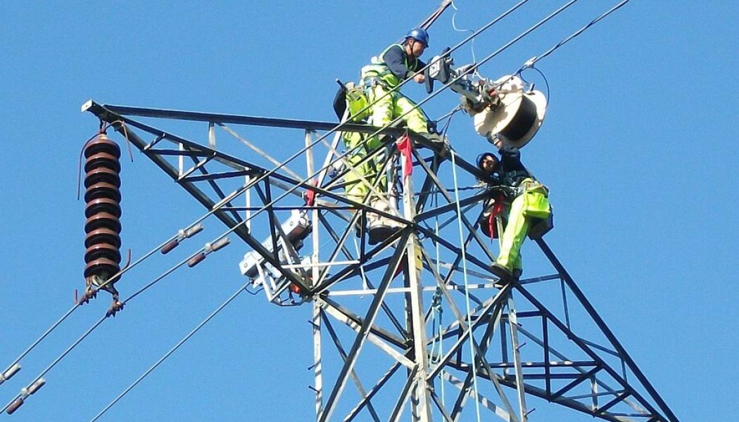Höghöjdsarbete pågår: Nya datakablar installeras på master. Högt uppe på en elektricitetsmast arbetar kablagearbetare med att lägga till en datakabel. De använder en specialmaskin som rullar upp kabeln på en trumma medan den färdas längs stödkabeln som sträcker sig mellan tornen. Maskinen, som drivs av egen motor, rör sig självständigt från ett torn till ett annat. När den når ett torn monteras den ner, flyttas över till andra sidan och fortsätter sedan sin färd. På bilden har drivmotorn redan flyttats över, men kabeltrumman väntar fortfarande på att hissas över tornet. Foto: Wikipedia Common, kredit: Sandpiper